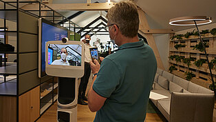 Ein Mann steht vor einem Telepräsenz-Roboter, auf dessen Bildschirm eine weitere Person zu sehen ist.