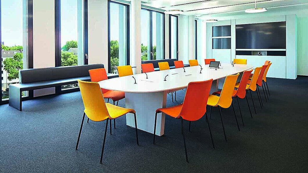 Konferenzraum im Gebäude des Handelsblatt mit einem großen Besprechungstisch mit orangenen Stühlen und einem Display an der Wand.
