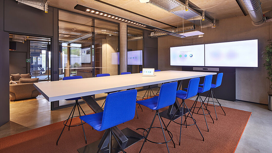 An einem langen Besprechungtisch stehen leuchtend blaue Stühle. Vor Kopf hängt ein sehr breiter Monitor für Videokonferenzen.