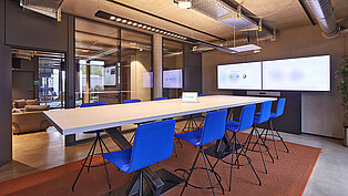 An einem langen Besprechungtisch stehen leuchtend blaue Stühle. Vor Kopf hängt ein sehr breiter Monitor für Videokonferenzen.