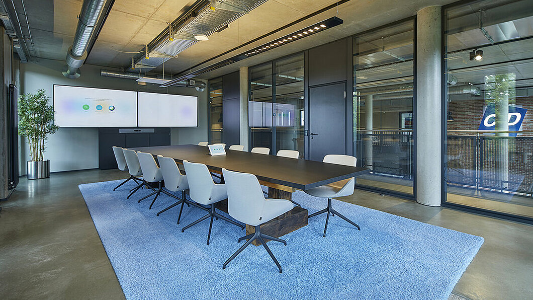 An einem langen Besprechungtisch stehen taubenblaue Stühle auf einem flauschigen Teppich in der selben Farbe. Vor Kopf hängt ein sehr breiter Monitor für Videokonferenzen.