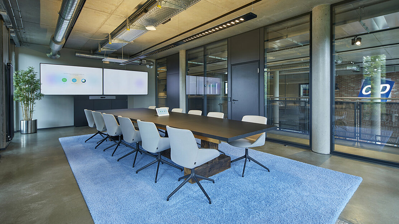 An einem langen Besprechungtisch stehen taubenblaue Stühle auf einem flauschigen Teppich in der selben Farbe. Vor Kopf hängt ein sehr breiter Monitor für Videokonferenzen.