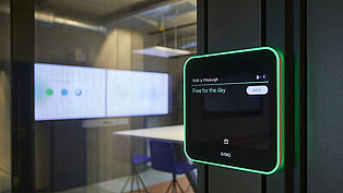 Eine elektronische Raumanzeige, die in grüner Schrift "Free For the day" zu lesen ist. Durch eine Glastür ist im Hintergrund der dazugehörige Konferenzraum zu sehen.