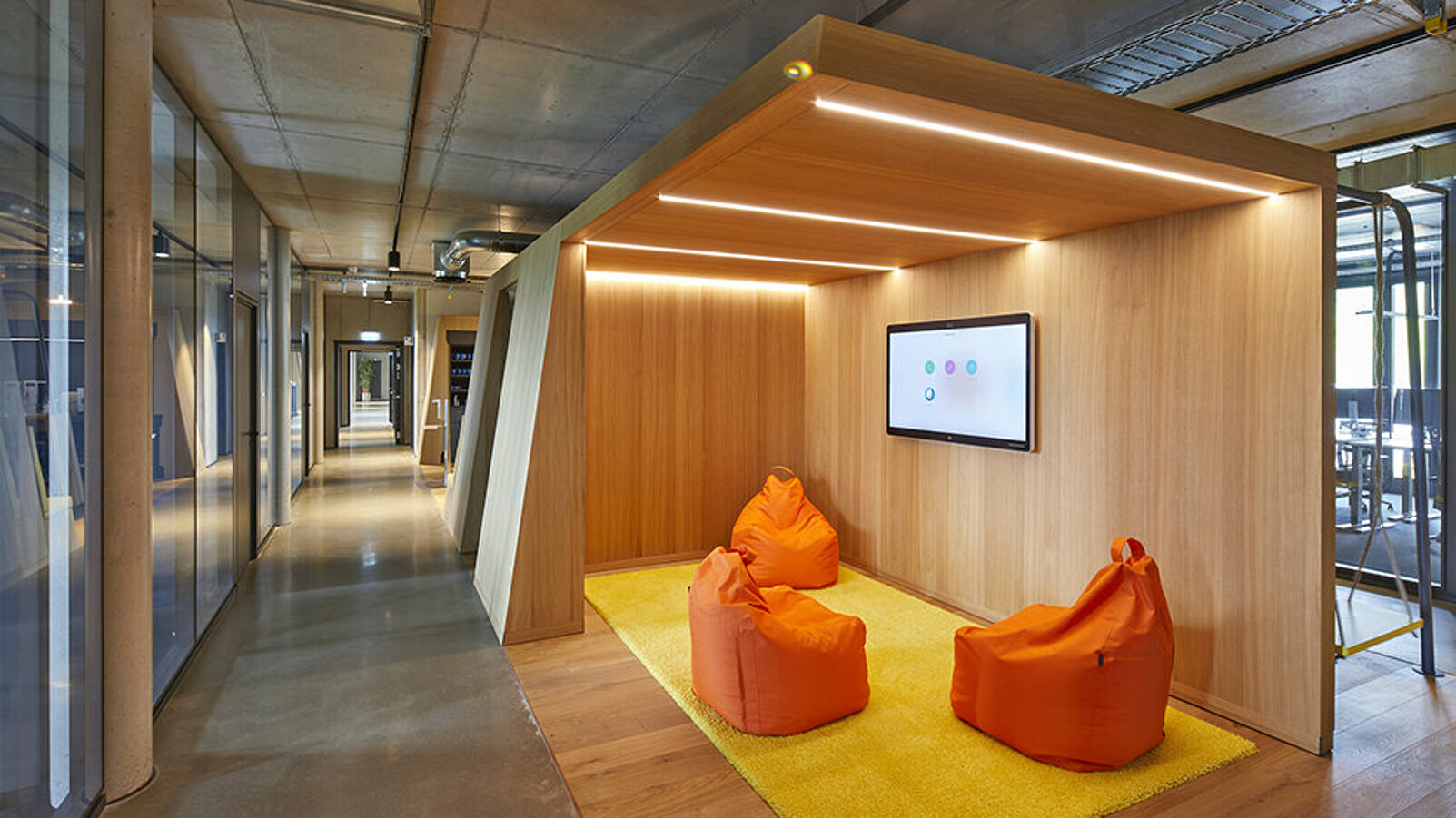 Drei orangefarbene Sitzsäcke auf einem flauschigen Teppich in einem kleinen, gemütlichen Besprechungsraum mit Monitor.