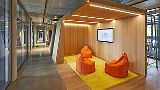 Drei orangefarbene Sitzsäcke auf einem flauschigen Teppich in einem kleinen, gemütlichen Besprechungsraum mit Monitor.